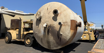 A large, dirty, off-white cylindrical industrial tank is being moved by a yellow forklift and another piece of heavy machinery, possibly a crane, in an outdoor industrial yard under a clear blue sky.
